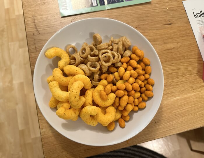 En tallrik med ostbågar, lökchips och jordnötter står på ett träbord.