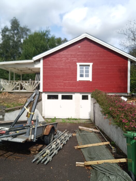 Röd husgavel med ny panel, 3-glasfönster och byggställning i förgrunden. Träplankor och täckt material ligger på marken framför.