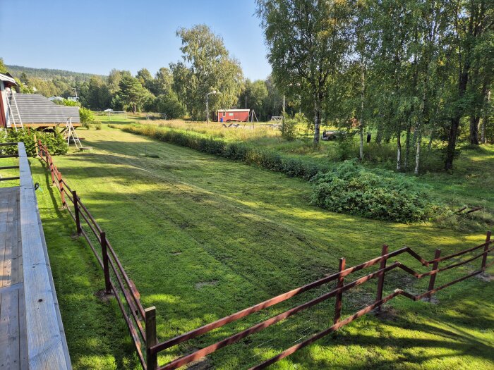 Nyklippt gräsmatta med utsikt över en omgivning med träd och ett rött hus i bakgrunden, sett från en veranda med ett trästaket.