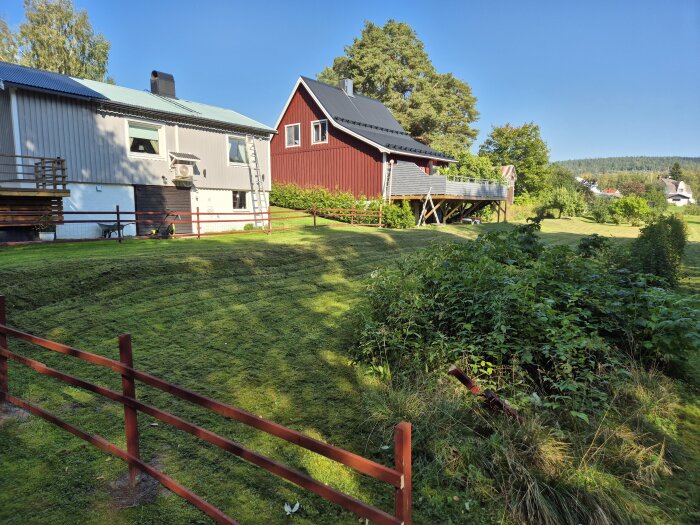 Två hus i ett grönt landskap med välklippt gräsmatta, ett grått och ett rött, omgivna av träd och buskar, under en klarblå himmel.