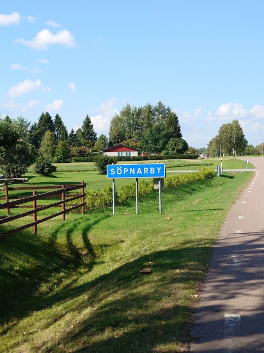 Vägskylt med texten "SÖPNARBY" vid landsväg, omgiven av grönska och en gård med ett rött tak i bakgrunden.