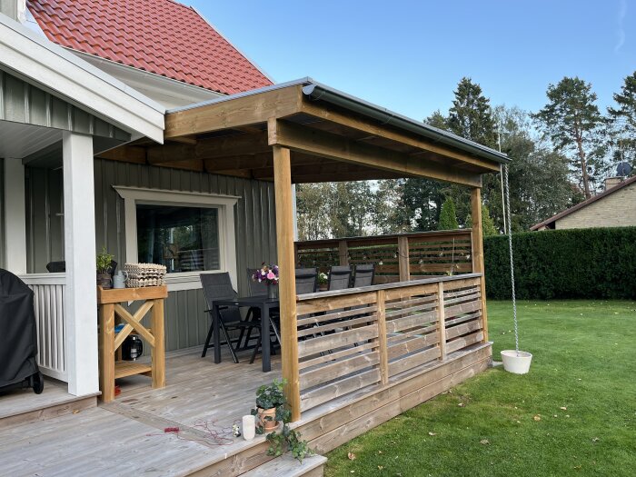 Altantak med hängränna och kedja monterad på ett hus. Utemöbler under taket och en kruka placerad vid foten av kedjan på verandan.