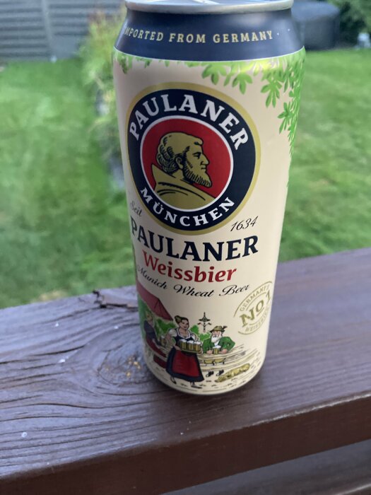 Burk med Paulaner Weissbier München Wheat Beer placerad på en brun träyta med suddig grönskande bakgrund.