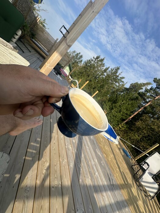 En hand håller en blå kaffekopp på en ny altan med trämöbler, omgiven av träd och blå himmel i bakgrunden.