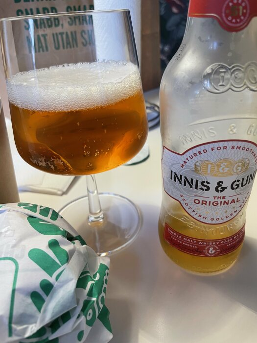 Närbild på en ölflaska av märket Innis & Gunn och ett glas öl bredvid ett inslaget paket som troligen innehåller en burgare.