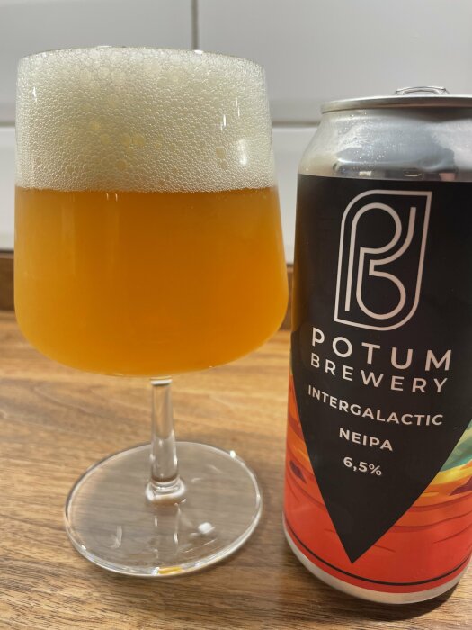 Glas med öl bredvid en burk Potum Brewery Intergalactic NEIPA 6,5% på ett träbord.