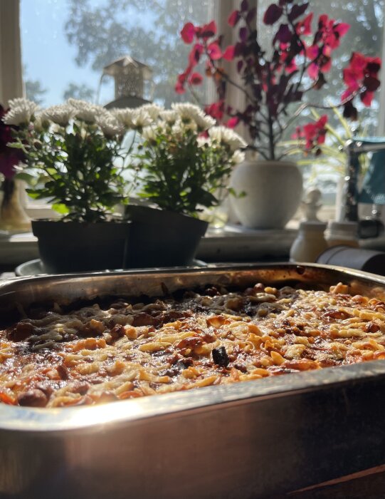 Makaronipudding i en ugnsform framför ett fönster, omgiven av vita och röda blommor.