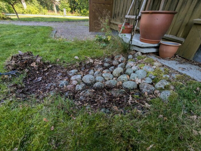 Stenfylld kallkälla på en gräsbevuxen tomt nära ett hus med krattade löv och skräp, omgiven av stora planteringskrukor och en kedja, i norra Västmanland.