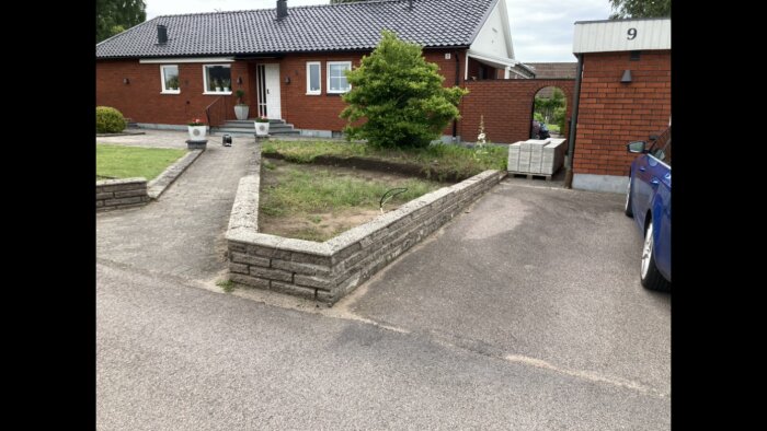 En enplans tegelvilla med svart takfot, förgård under renovering, låga murar och nyplanterad häck. Stenplattor travade intill garageinfart.