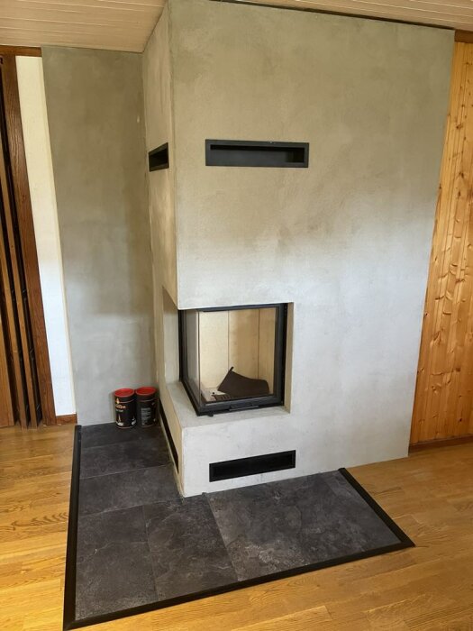 Bild av en nyinstallerad kamin med ojämn puts på väggen runt om. Mörka plattor runt kaminen, två burkar bredvid. Väggpanelen till höger är av trä.