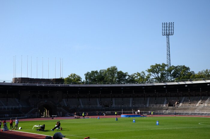 Stockholms stadion en solig dag med få besökare vid atletiska aktiviteter på planen, tomma läktare i bakgrunden och ett torn med strålkastare synligt.