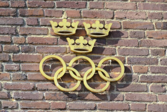 Tre gyllene kronor och de olympiska ringarna i guld monterade på en tegelvägg.