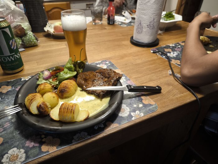 En tallrik med grillad entrecote, hasselbackspotatis, sås och sallad på ett bord med en öl, servett och matlagningstillbehör.