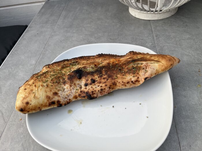 En hembakad calzone ligger på en vit tallrik, dekorerad med örtkryddor på ett grått bord.