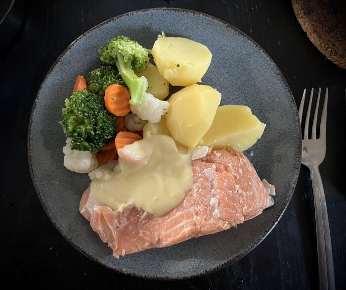 Tallrik med alspånsrökt lax, täckt med hollandaisesås, serverat med kokt potatis och en blandning av kokta grönsaker som broccoli, morötter och blomkål.