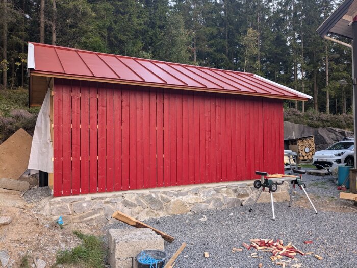 Röd byggnad med nyinstallerad målade paneler, tak och grundarbeten av sten synliga. Verktyg och material utspridda på marken. Skog i bakgrunden.