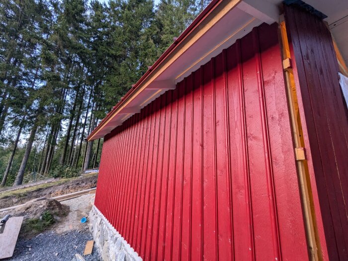 Rödmålad fasad på ett hus med träd i bakgrunden. Lockläkt spikade och vitmålad ribba upptill under takfoten. Byggmaterial på marken.