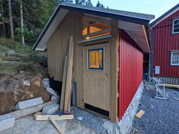 Halvfärdigt förråd med grundmålad träpanel och liten dörr monterad på framsidan. Stöttande material och virke ligger vid sidan av förrådet.