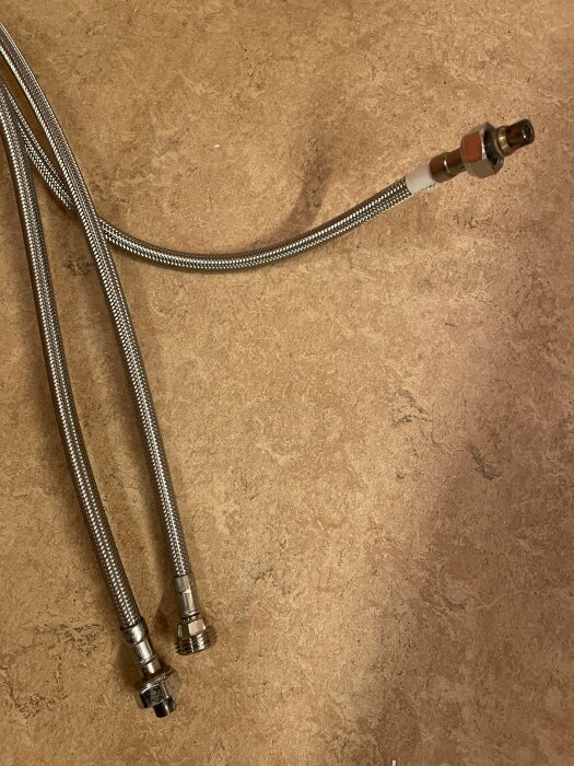 Två metallomspunna slangar med kopplingar som ligger på ett beige golv, en koppling stickar ut från ena änden av en slang.