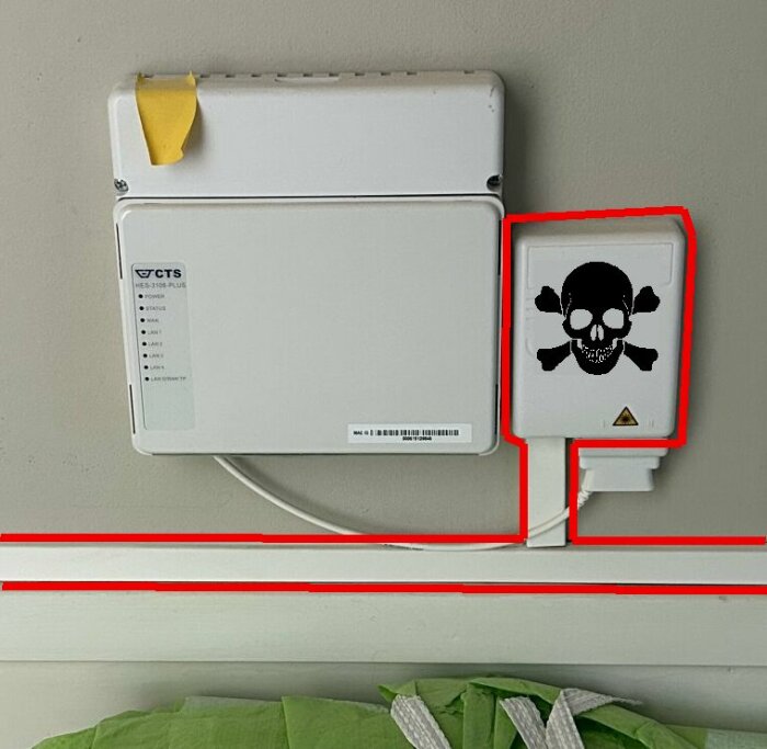 En HES-3106-PLUS switch med VLAN fäst på en vägg bredvid en liten vit låda märkt med en dödskalle-inte underhållen och rödmarkerade områden.