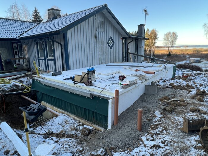 Byggarbetsplats med isolerad grund och byggmaterial utanför ett hus under renovering i vintrig miljö. Verktyg och material ligger på grunden.