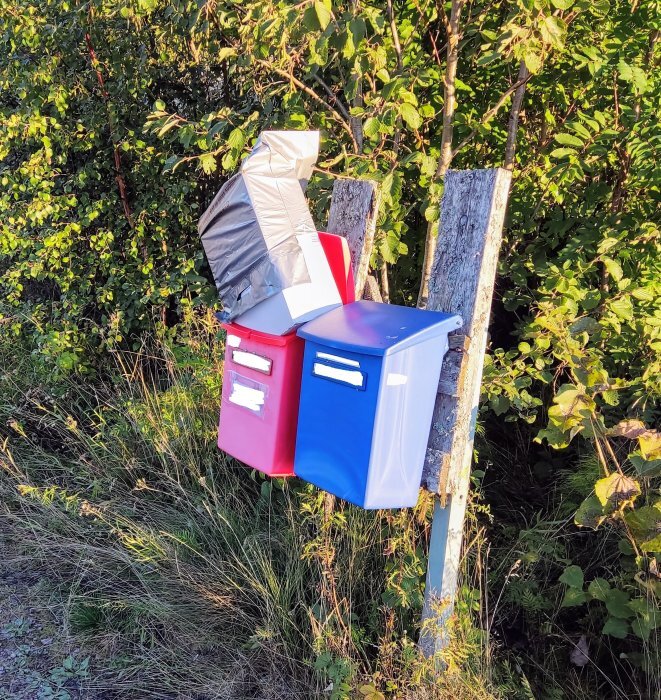 Två brevlådor, en röd och en blå, med paket som inte får plats i brevlådorna, placerade vid en lantlig väg.