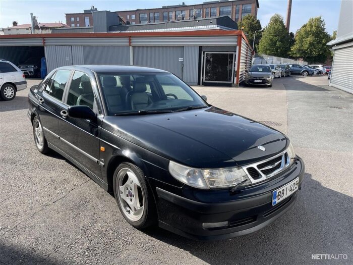 Svart Saab 9-5 Aero från 1999 parkerad på en asfaltsplan framför en byggnad.