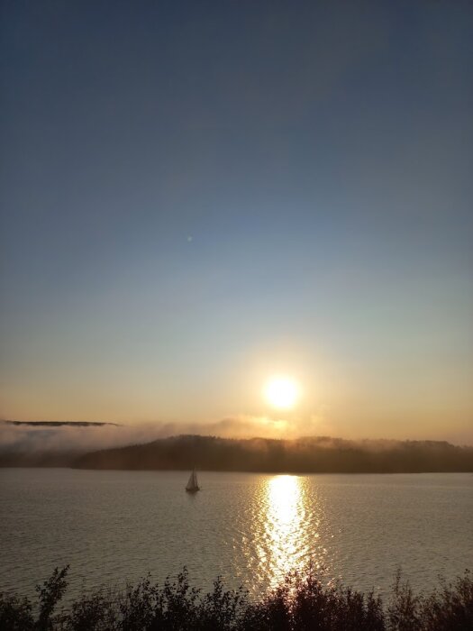 Segelbåt på en sjö i solnedgången med dimma över skogen i bakgrunden.