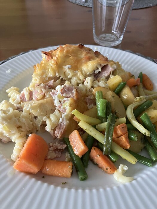 Tallrik med makaronipudding med purjolök och pastrami samt ugnsrostade rotfrukter, inklusive morötter och gröna bönor, stående på ett bord.