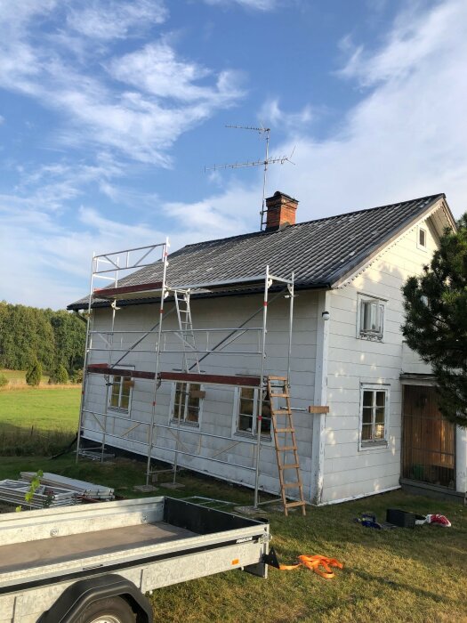 Tvåvåningshus med byggställning och stege vid fasaden, skorsten och TV-antenn på taket. Trailer och verktyg synliga i förgrunden.