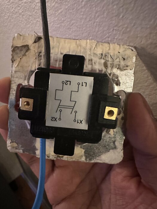 Närbild på en elektronisk komponent med anslutna kablar och en kopplingsschema-etikett på framsidan, hålles i en hand mot en vägg.