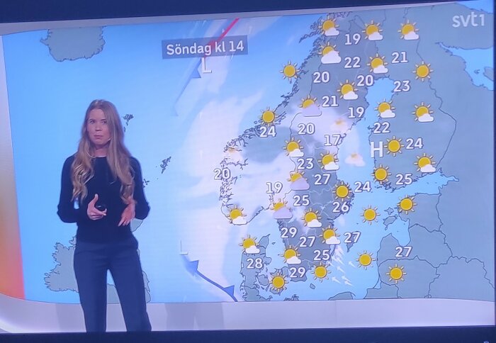 Väderpresentatör från SVT visar en väderprognos för Skandinavien med temperaturer och symboler för sol och moln på en lördag kväll.