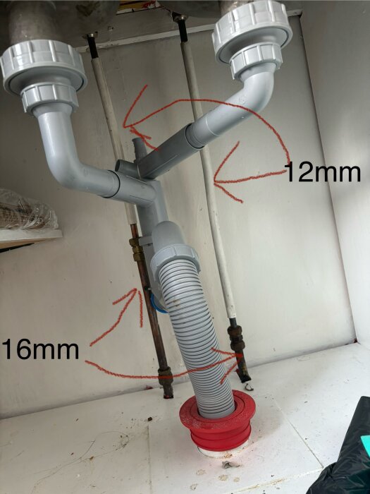 Avloppsrör och vattenrör under en diskbänk; två vertikala rör markerade med texten "12mm" och "16mm", förberedelse inför byte av köksblandare.