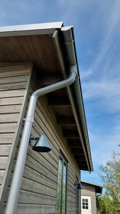 Träpanel, takränna och utomhuslampa på ett hus nära havet. Förrådsdörr och växtlighet i bakgrunden.