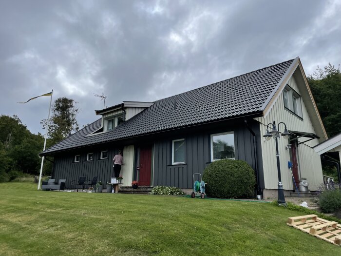 En villa med mörkgrå fasad, större fönster och sluttande tak. En person står på en stege vid husets entré och arbetar på en dörr under molnig himmel.