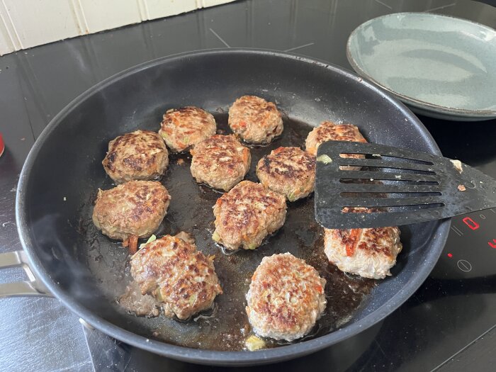 Stekta köttfärsbiffar med grönsaker i en stekpanna, en spatel bredvid dem på en spis.