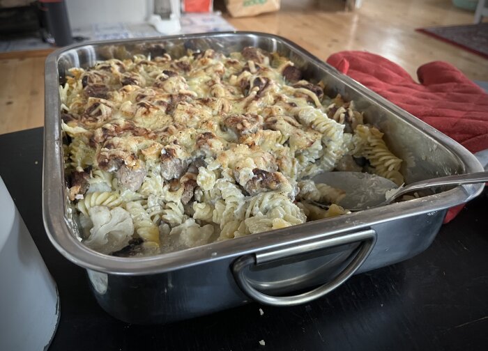 Gratäng med makaroner, ost och kött i en rektangulär ugnsform på ett bord, en röd grytvante bredvid.