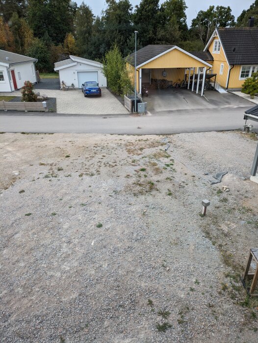 En grusad yta med spridda ogräs, bredvid en villaväg med hus och parkeringar.