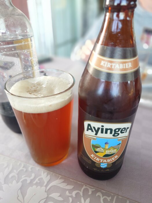 En ölflaska märkt Ayinger Kirtabier och ett fyllt ölglas stående på en dukad yta.