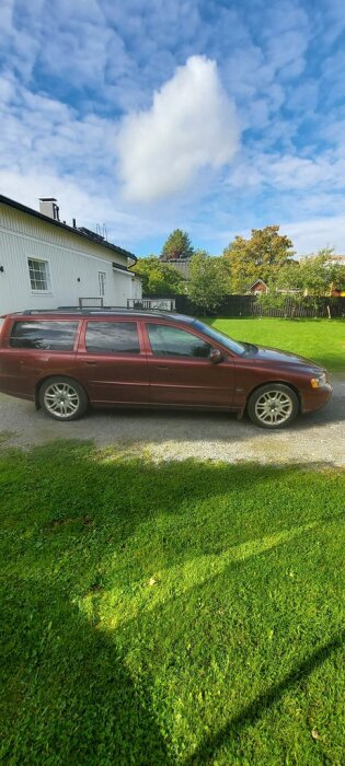 Röd kombibil parkerad på grusväg framför vit byggnad.