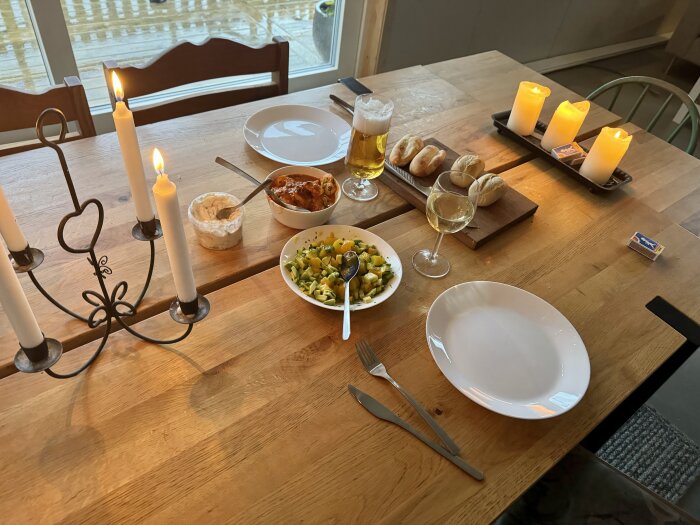 En dukad träbord med stekta räkor, sallad, bröd, ett glas öl och ett glas vin, tända ljus, två tomma tallrikar och bestick.