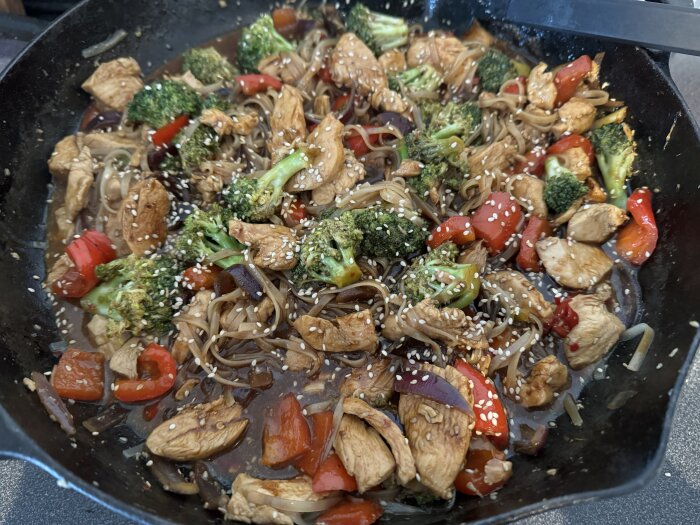 Kycklingwok med broccoli, röd paprika, nudlar och sesamfrön lagad i en stekhäll.