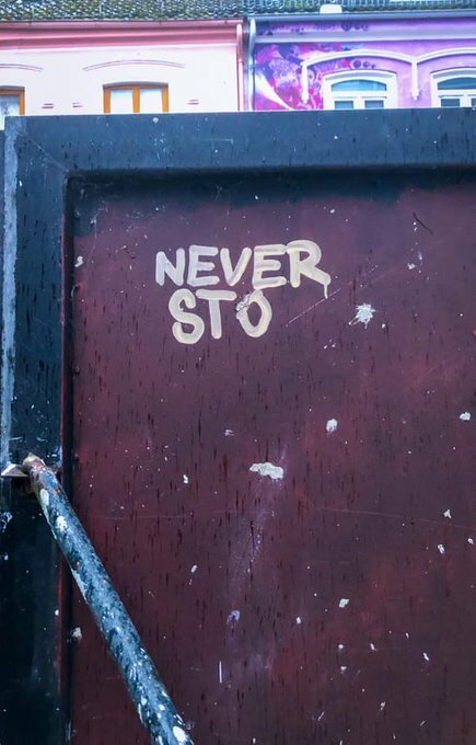 Klistermärke på vägg som saknar bokstäver, med texten "NEVER STO", framför färgglatt hus.