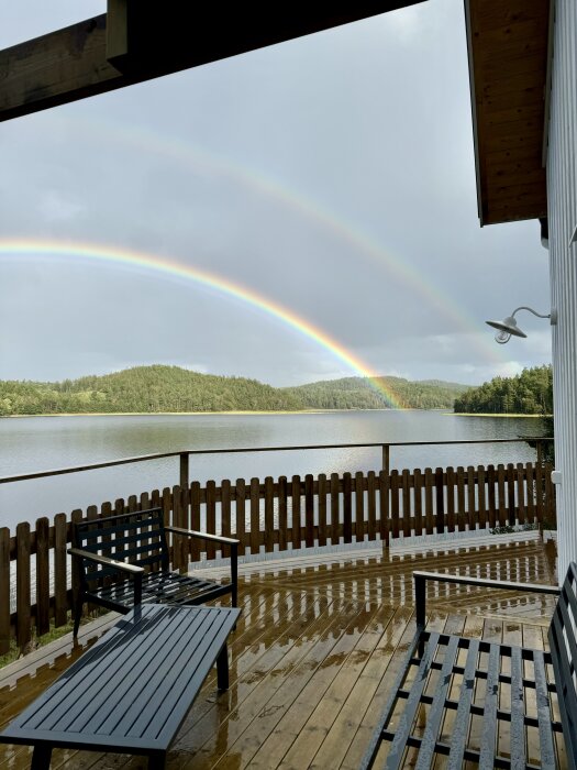 Trädäck med bänk vid sjö, dubbel regnbåge synlig på himlen, omgiven av grönska och staket med utsikt över vattnet.