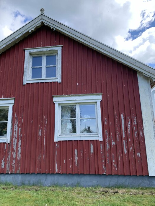 Röd timmerstuga med vitt fönsterfoder och avskalad färg på fasaden, grå sockel, tre-fönster på fasaden, grönt gräs nedanför och molnig himmel ovanför.