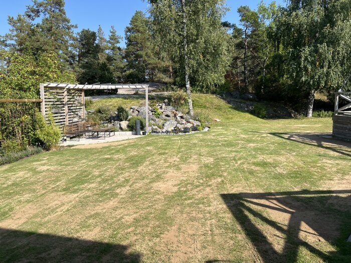 Nyligen renoverad gräsmatta med lågklippt gräs, synliga låga punkter, paviljong i bakgrunden och omgivande vegetation under en klarblå himmel.