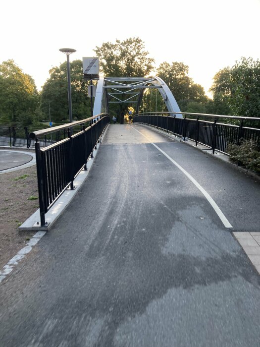 Gång- och cykelbro med svarta räcken och en upplyst lampa, omgiven av träd, vid solnedgång.