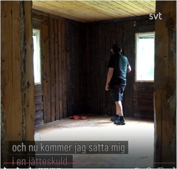 En person står i ett rum med ljus träpanel i taket och mörka träväggar, medan text från ett tv-program visas över bilden.