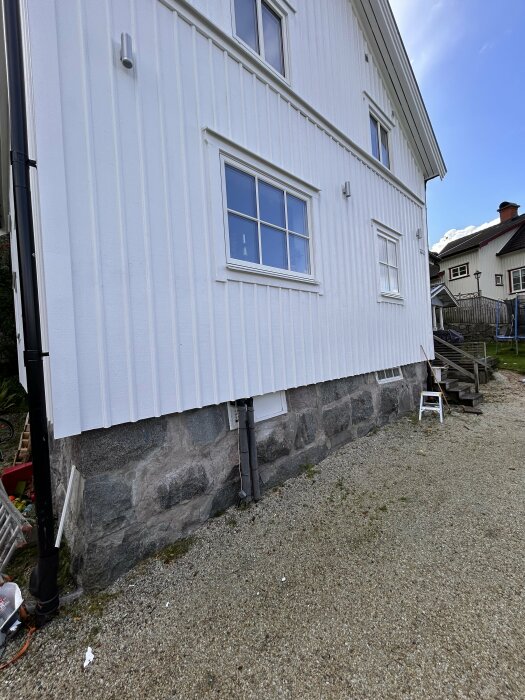 Vitmålad träpanel på husets vägg, med oregelbundna granitblock i husgrunden. Markytan består av grus och en liten stege står intill väggen.