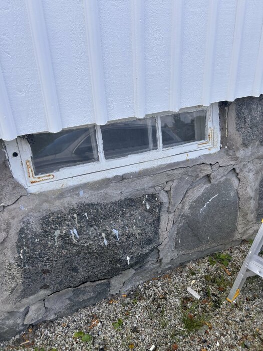 Närbild på husgrund av ojämna granitblock och del av vitmålad fasadpanel ovanför. Ett fönster med rostig ram är synligt.